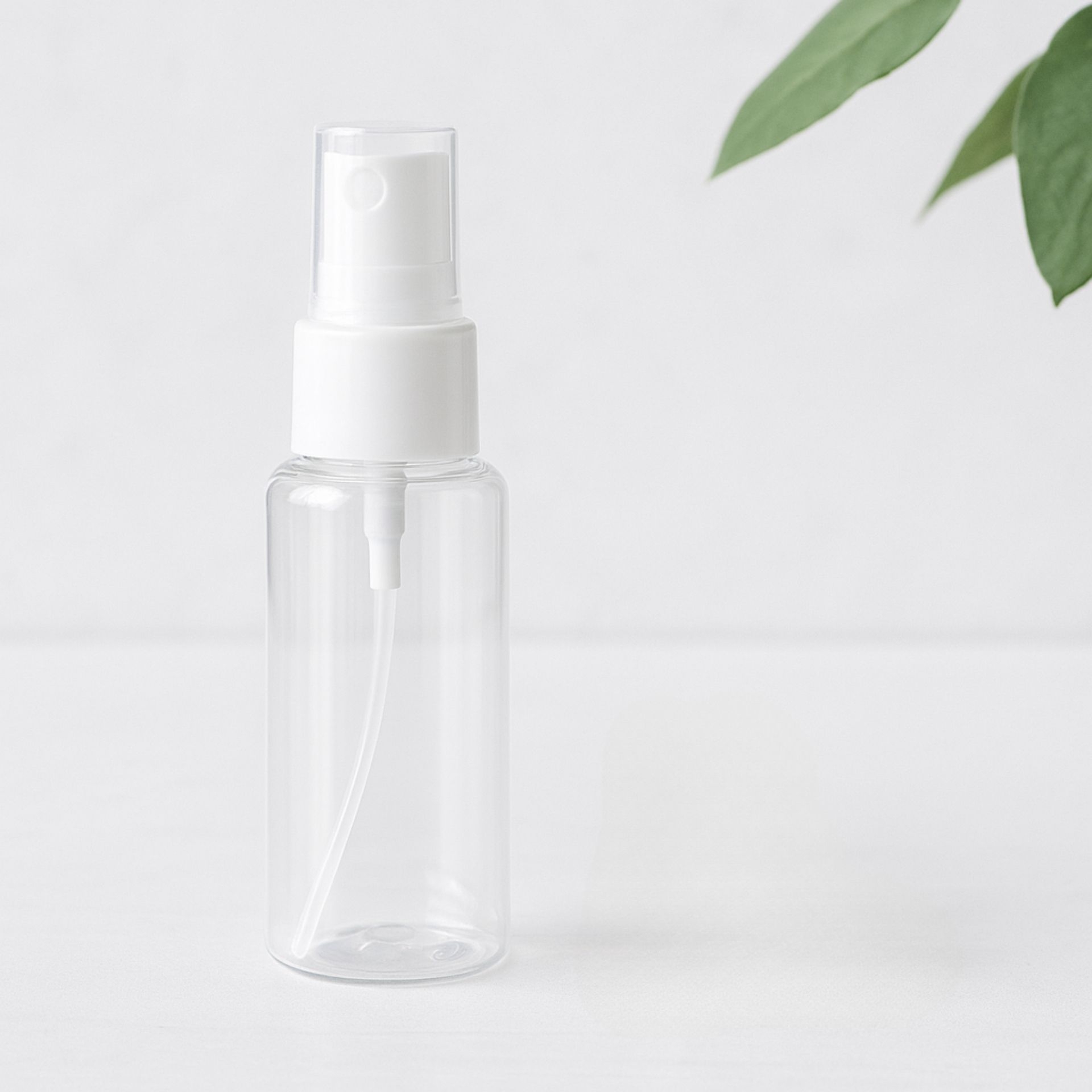 Transparente 50ml Sprayflasche mit weissem Spruehkopf_ minimalistisch auf hellem Hintergrund mit Blattdeko fotografiert