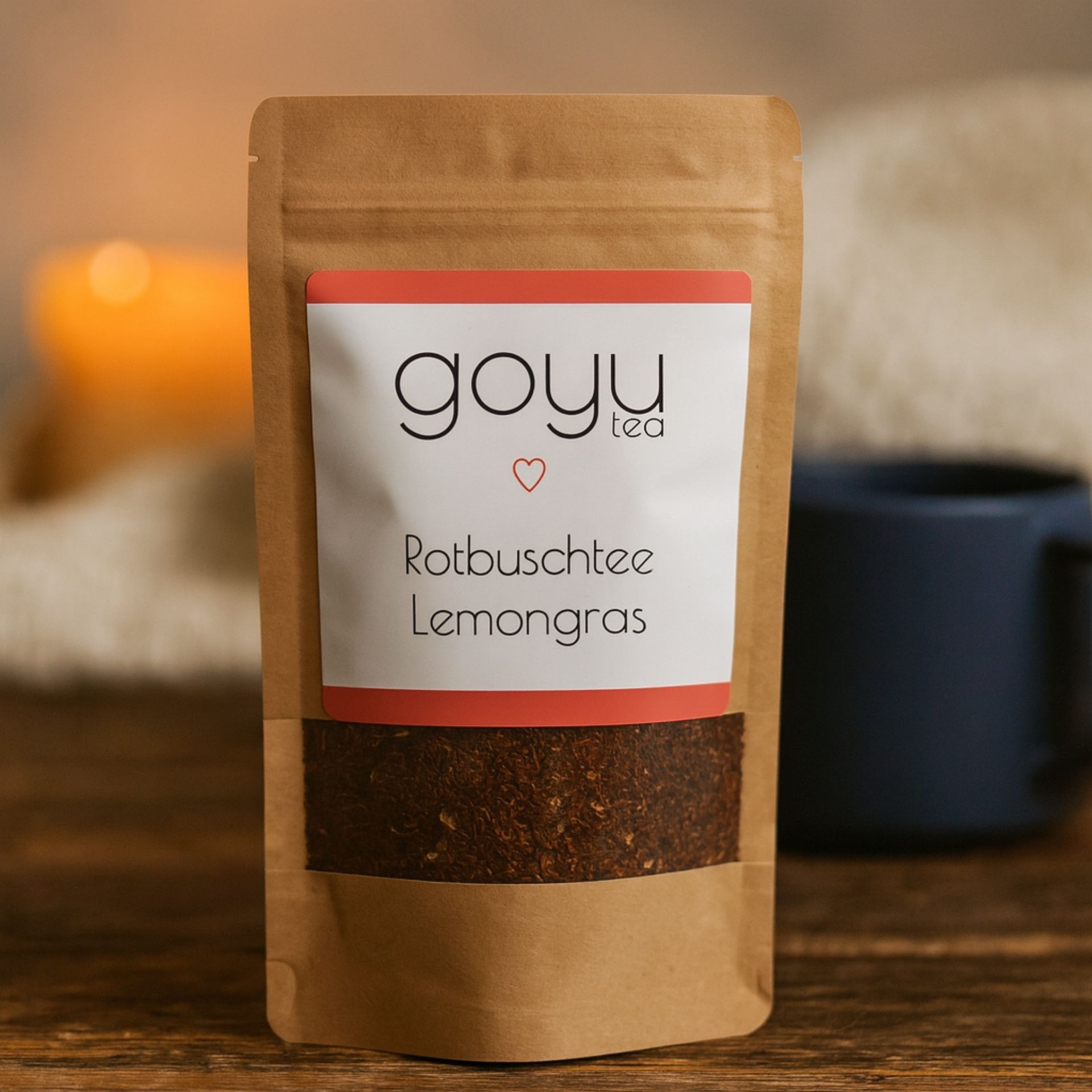 Braune Teeverpackung „Rotbuschtee Lemongras“ von goyu tea auf Holztisch_ im Hintergrund eine dunkle Tasse und Kerzenschein