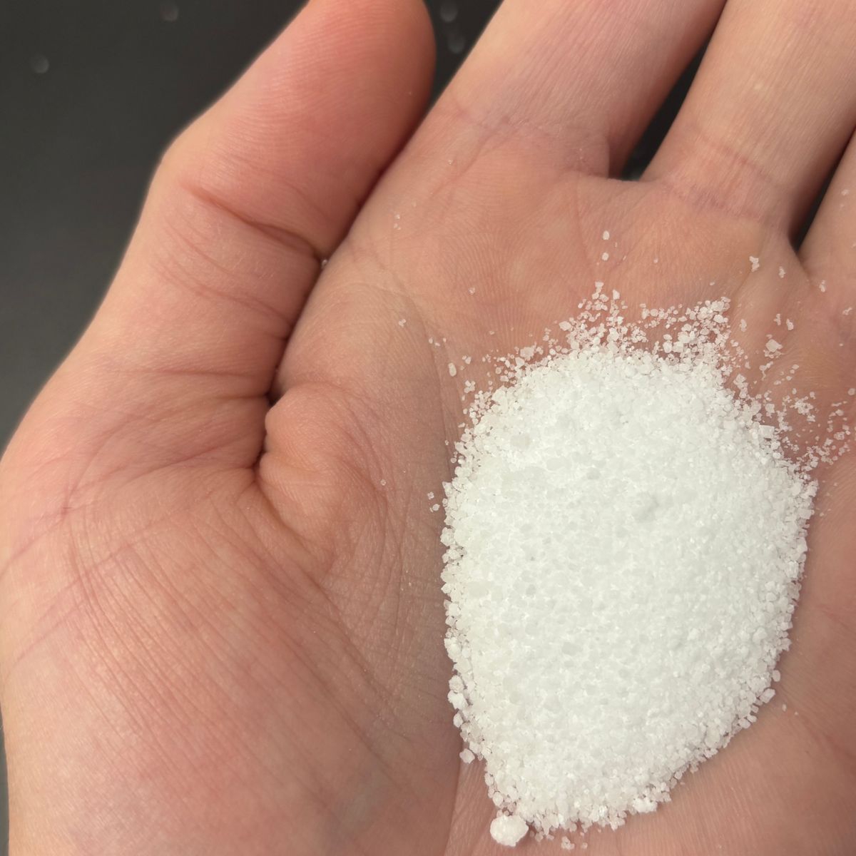 Hand haelt weisse Calciumchlorid-Kristalle ueber dunklem Hintergrund