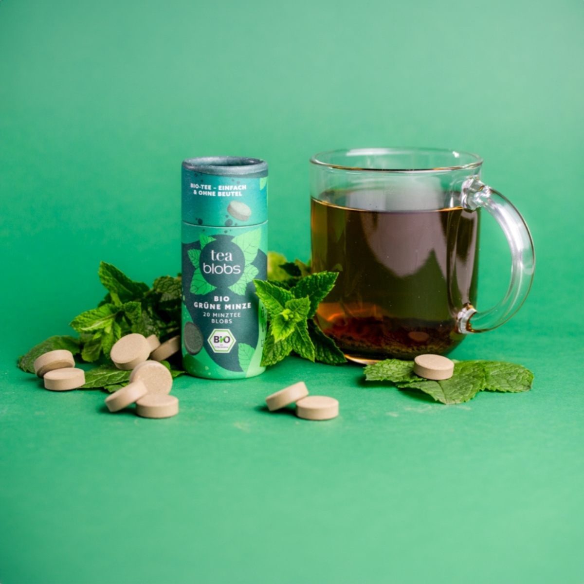 Teablobs Bio Gruene Minze mit losem Tee_ Blobs_ Glas Minztee und Minzblaettern auf gruenem Hintergrund