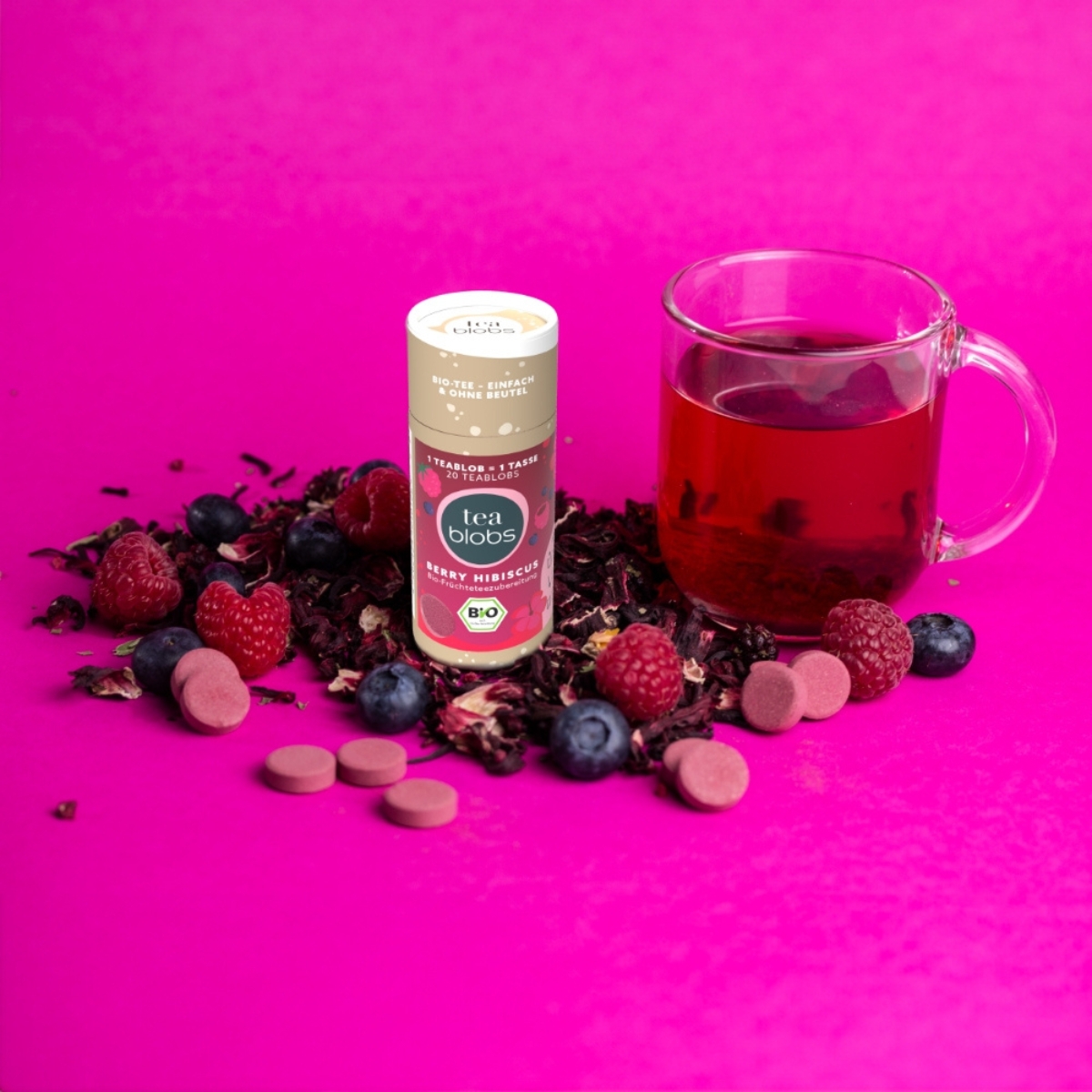 Teablobs Berry Hibiscus Dose mit Beeren_ Hibiskusblueten und Glas roten Tees auf pinkem Hintergrund