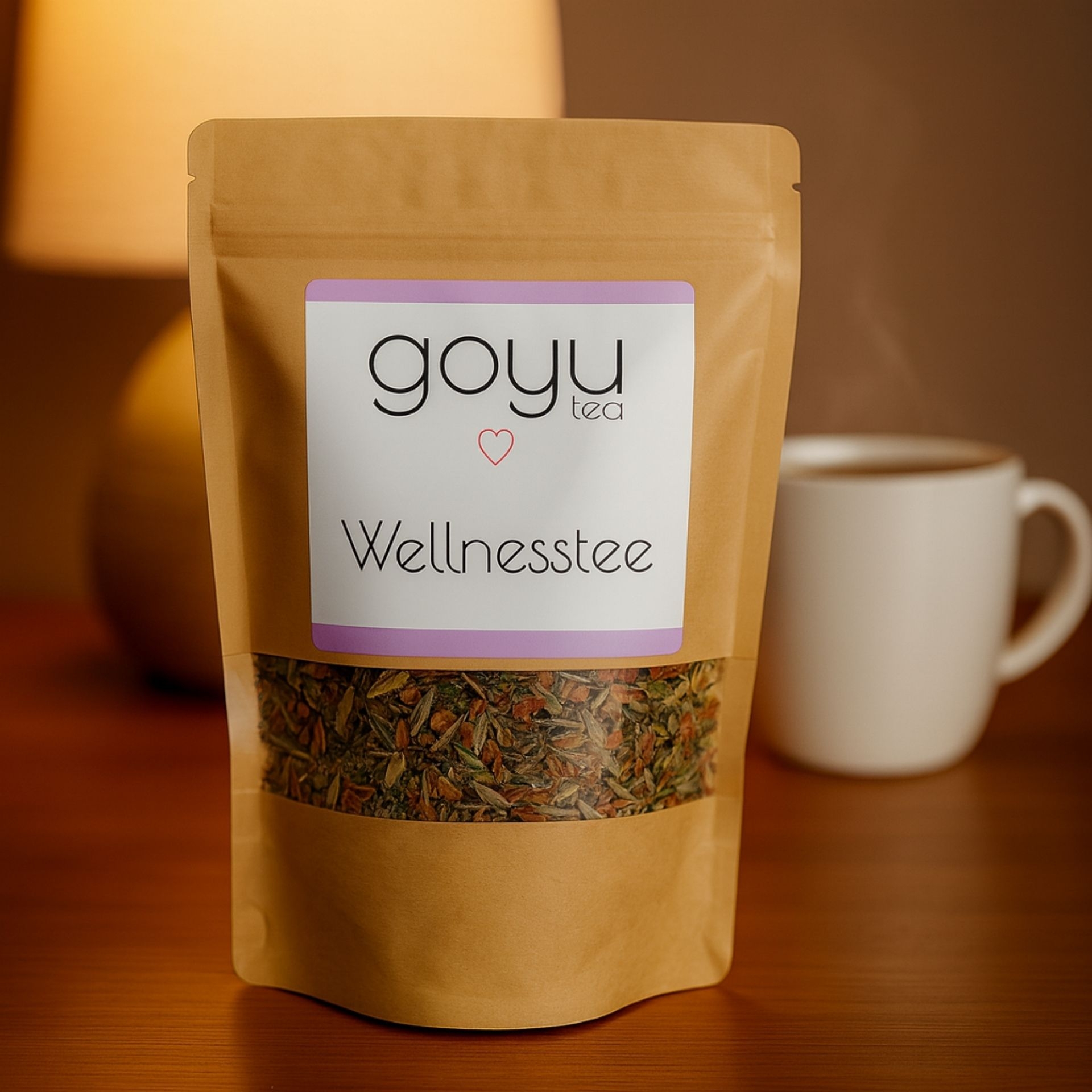 Beutel „Wellnesstee“ von goyu tea auf Holztisch_ im Hintergrund eine dampfende weisse Tasse und eine warme Tischlampe