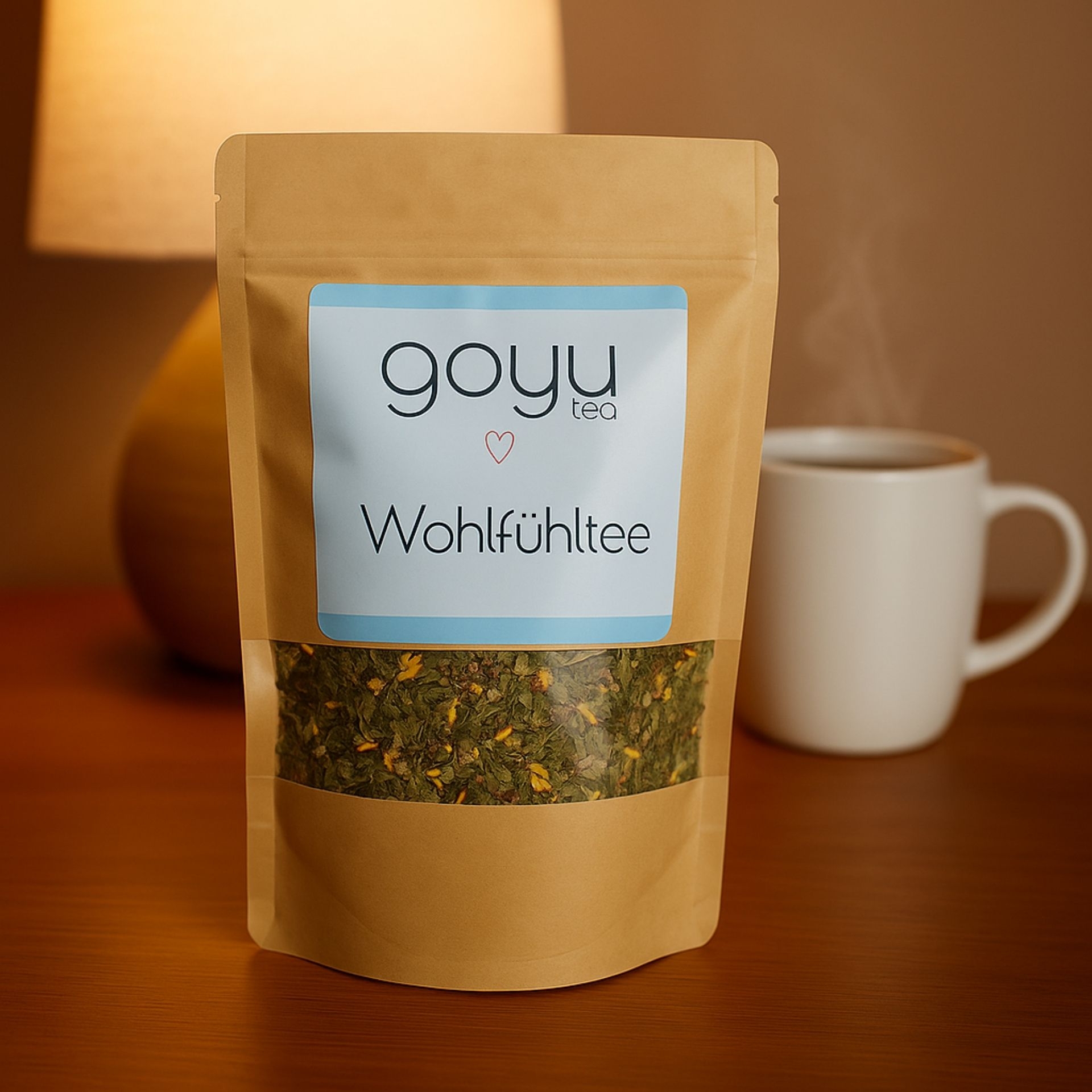 Beutel „Wohlfuehltee“ von goyu tea auf Holztisch_ im Hintergrund eine weisse Tasse mit dampfendem Tee und eine Lampe